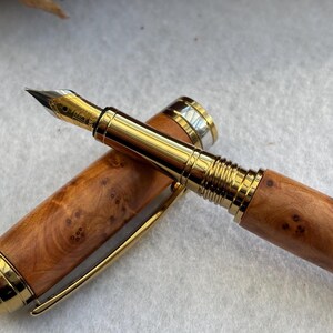 Puede incluir: Una pluma estilogr&aacute;fica de lujo con un cuerpo de madera y detalles dorados. La tapa de la pluma est&aacute; quitada, revelando el plum&iacute;n y el intrincado patr&oacute;n de vetas de la madera. La pluma est&aacute; sobre una superficie blanca.