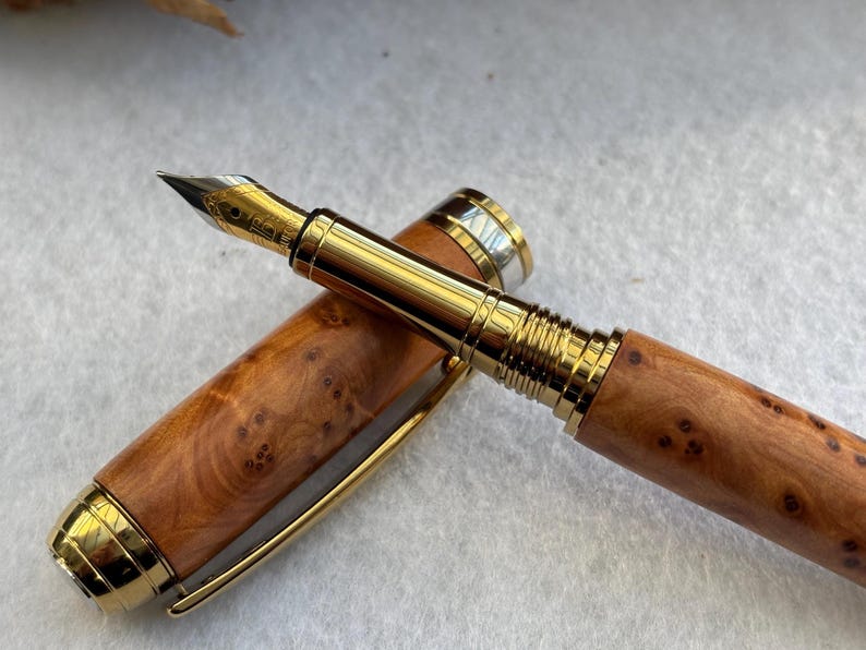 Puede incluir: Una pluma estilogr&aacute;fica de lujo con un cuerpo de madera y detalles dorados. La tapa de la pluma est&aacute; quitada, revelando el plum&iacute;n y el intrincado patr&oacute;n de vetas de la madera. La pluma est&aacute; sobre una superficie blanca.