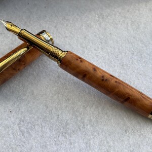 Puede incluir: Una pluma estilogr&aacute;fica de lujo con plum&iacute;n y detalles dorados. El cuerpo de la pluma es de madera marr&oacute;n claro con manchas oscuras, y la tapa est&aacute; abierta, revelando el plum&iacute;n dorado. Es un instrumento de escritura.