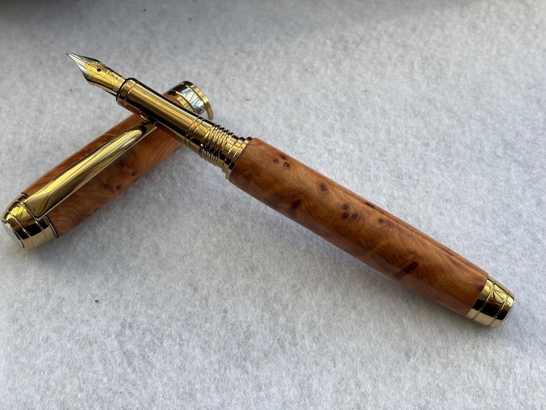 Puede incluir: Una pluma estilogr&aacute;fica de lujo con plum&iacute;n y detalles dorados. El cuerpo de la pluma es de madera marr&oacute;n claro con manchas oscuras, y la tapa est&aacute; abierta, revelando el plum&iacute;n dorado. Es un instrumento de escritura.
