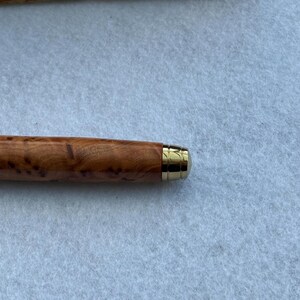 Puede incluir: Primer plano de un bol&iacute;grafo de madera con punta de metal dorada. El cuerpo del bol&iacute;grafo es de color marr&oacute;n claro con un patr&oacute;n de vetas de madera &uacute;nico. La punta dorada a&ntilde;ade un toque de elegancia.