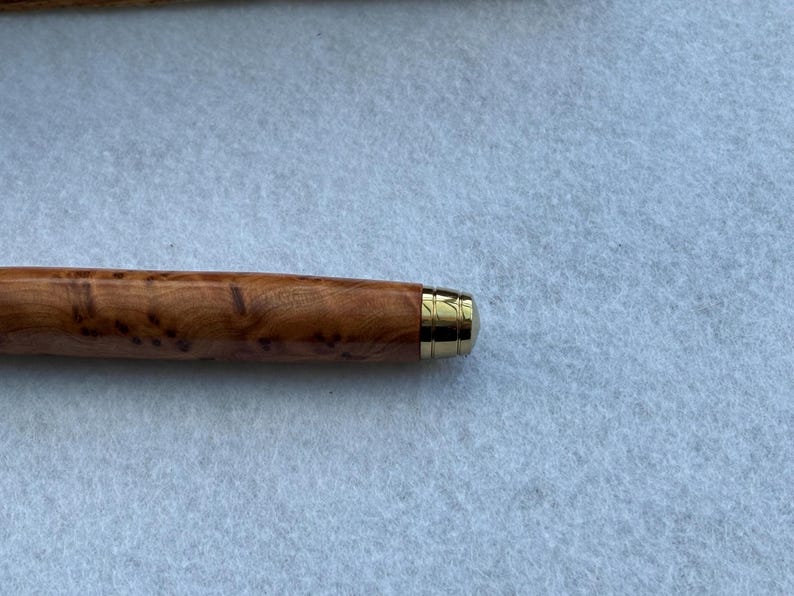 Puede incluir: Primer plano de un bol&iacute;grafo de madera con punta de metal dorada. El cuerpo del bol&iacute;grafo es de color marr&oacute;n claro con un patr&oacute;n de vetas de madera &uacute;nico. La punta dorada a&ntilde;ade un toque de elegancia.