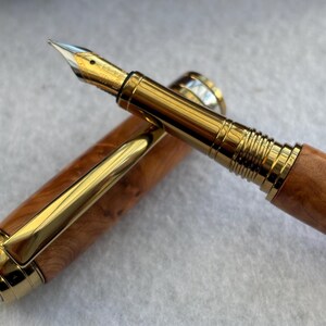 Puede incluir: Una pluma estilogr&aacute;fica de lujo con plum&iacute;n y detalles dorados. El cuerpo de la pluma es de madera marr&oacute;n claro con un patr&oacute;n de vetas natural. El capuch&oacute;n est&aacute; quitado, revelando la punta de escritura. Un instrumento de escritura cl&aacute;sico.