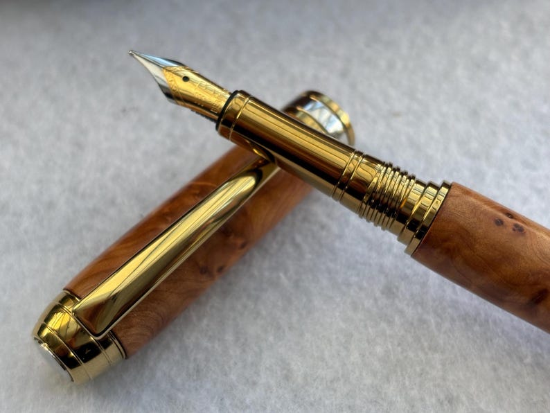 Puede incluir: Una pluma estilogr&aacute;fica de lujo con plum&iacute;n y detalles dorados. El cuerpo de la pluma es de madera marr&oacute;n claro con un patr&oacute;n de vetas natural. El capuch&oacute;n est&aacute; quitado, revelando la punta de escritura. Un instrumento de escritura cl&aacute;sico.