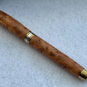 Puede incluir: Pluma estilogr&aacute;fica de madera con un patr&oacute;n de vetas de madera natural. El bol&iacute;grafo tiene detalles dorados en la punta, la tapa y una banda en el centro. Es un instrumento de escritura.