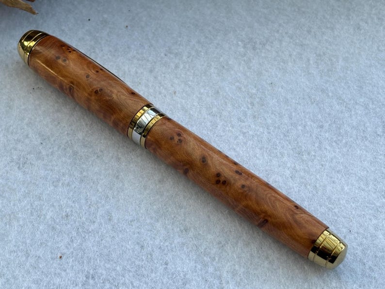 Puede incluir: Pluma estilogr&aacute;fica de madera con un patr&oacute;n de vetas de madera natural. El bol&iacute;grafo tiene detalles dorados en la punta, la tapa y una banda en el centro. Es un instrumento de escritura.