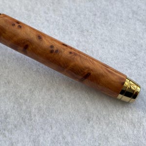 Puede incluir: Primer plano de un bol&iacute;grafo de madera con acabado marr&oacute;n claro y manchas marrones oscuras. El bol&iacute;grafo tiene detalles met&aacute;licos dorados en la punta y en la parte superior. El bol&iacute;grafo est&aacute; sobre una superficie blanca.