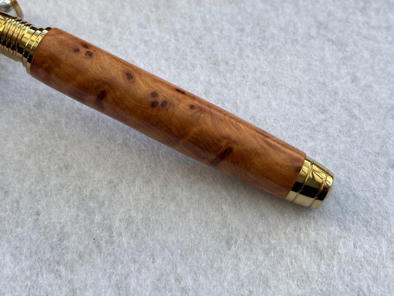 Puede incluir: Primer plano de un bol&iacute;grafo de madera con acabado marr&oacute;n claro y manchas marrones oscuras. El bol&iacute;grafo tiene detalles met&aacute;licos dorados en la punta y en la parte superior. El bol&iacute;grafo est&aacute; sobre una superficie blanca.