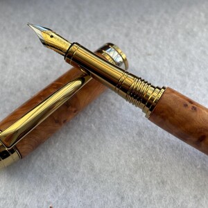 Puede incluir: Una pluma estilogr&aacute;fica de lujo con plum&iacute;n y detalles dorados. El cuerpo de la pluma es de madera con un patr&oacute;n de vetas natural. El capuch&oacute;n est&aacute; quitado, revelando el plum&iacute;n y el clip dorado.