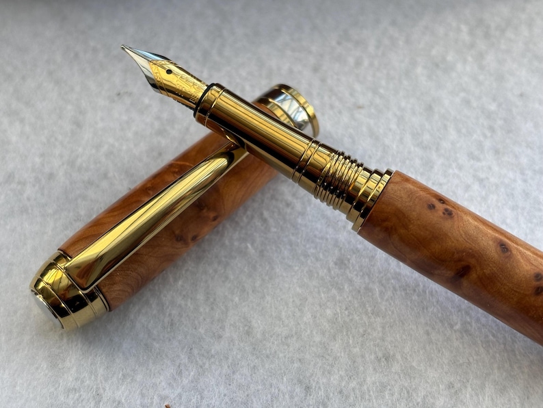 Puede incluir: Una pluma estilogr&aacute;fica de lujo con plum&iacute;n y detalles dorados. El cuerpo de la pluma es de madera con un patr&oacute;n de vetas natural. El capuch&oacute;n est&aacute; quitado, revelando el plum&iacute;n y el clip dorado.