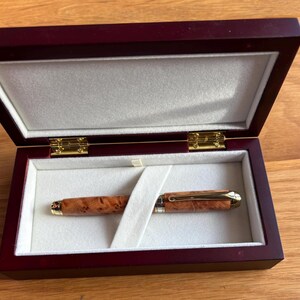 Puede incluir: Un bol&iacute;grafo de madera con clip y detalles dorados descansa dentro de una caja de madera burdeos abierta, con interior blanco. El bol&iacute;grafo presenta un patr&oacute;n de vetas de madera natural. La caja est&aacute; abierta, revelando el bol&iacute;grafo.