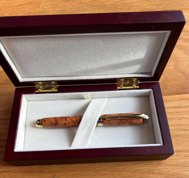 Puede incluir: Un bol&iacute;grafo de madera con clip y detalles dorados descansa dentro de una caja de madera burdeos abierta, con interior blanco. El bol&iacute;grafo presenta un patr&oacute;n de vetas de madera natural. La caja est&aacute; abierta, revelando el bol&iacute;grafo.