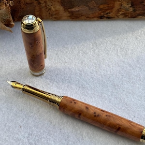 Puede incluir: Una pluma estilogr&aacute;fica de madera con detalles dorados. El capuch&oacute;n est&aacute; quitado, revelando el plum&iacute;n. El bol&iacute;grafo tiene un patr&oacute;n de vetas de madera natural. Es un instrumento de escritura.