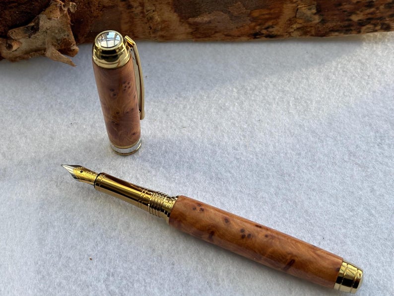 Puede incluir: Una pluma estilogr&aacute;fica de madera con detalles dorados. El capuch&oacute;n est&aacute; quitado, revelando el plum&iacute;n. El bol&iacute;grafo tiene un patr&oacute;n de vetas de madera natural. Es un instrumento de escritura.