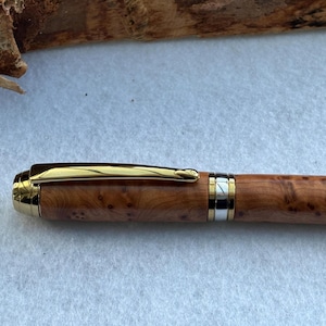 Puede incluir: Un bol&iacute;grafo de madera con clip y detalles dorados. El cuerpo del bol&iacute;grafo presenta un patr&oacute;n de vetas de madera natural con una banda dorada y plateada cerca de la punta. El bol&iacute;grafo est&aacute; sobre una superficie blanca.