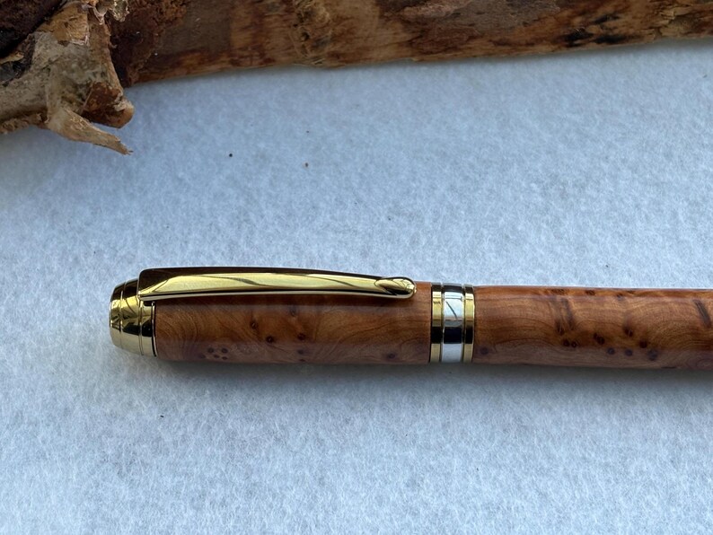 Puede incluir: Un bol&iacute;grafo de madera con clip y detalles dorados. El cuerpo del bol&iacute;grafo presenta un patr&oacute;n de vetas de madera natural con una banda dorada y plateada cerca de la punta. El bol&iacute;grafo est&aacute; sobre una superficie blanca.