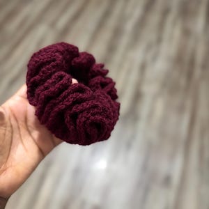 May include: A burgundy crocheted scrunchie, a hair accessory, is held in a hand. The scrunchie is made of a thick, textured yarn, and the color is a deep, rich shade of red-violet. The scrunchie is round and gathered, with visible stitching.