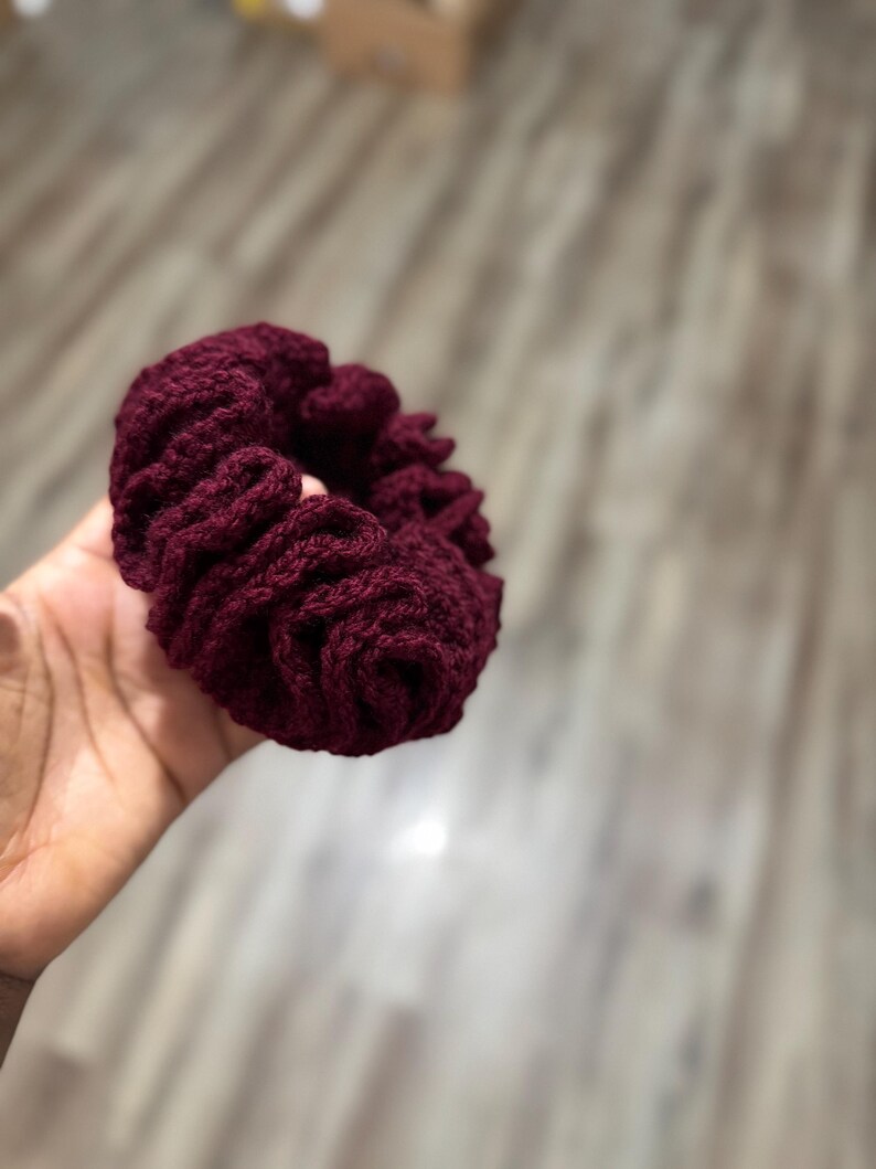 May include: A burgundy crocheted scrunchie, a hair accessory, is held in a hand. The scrunchie is made of a thick, textured yarn, and the color is a deep, rich shade of red-violet. The scrunchie is round and gathered, with visible stitching.