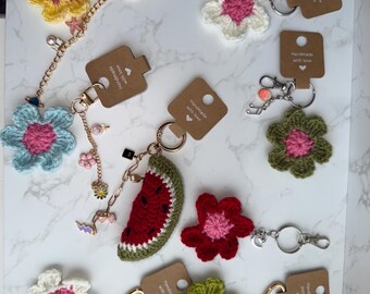 May include: A collection of handmade crochet keychains featuring various flower shapes in yellow, white, light blue, olive green, and red, along with a watermelon slice design. Each keychain includes a metal ring and a brown tag labeled Handmade with love.