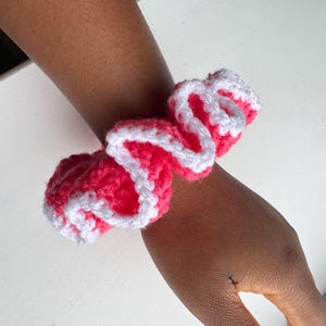 May include: A handmade crochet scrunchie in bright pink with a white trim, wrapped around a wrist. The scrunchie is made of soft yarn and has a ruffled appearance. This hair accessory adds a pop of color.
