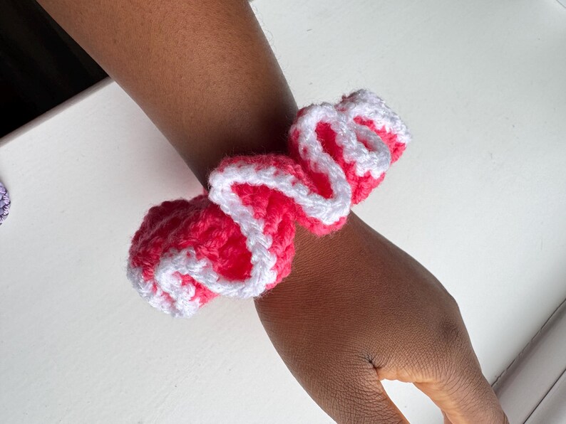 May include: A handmade crochet scrunchie in bright pink with a white trim, wrapped around a wrist. The scrunchie is made of soft yarn and has a ruffled appearance. This hair accessory adds a pop of color.