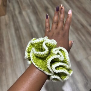 May include: A handmade scrunchie in a ruffled design. The scrunchie features a vibrant green color with a white trim, creating a contrasting and textured appearance. The accessory is designed to hold hair in place.