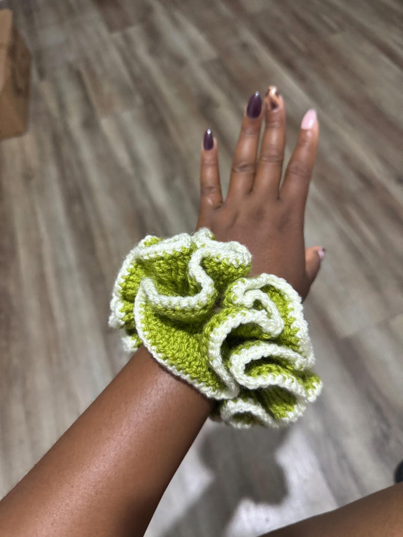 May include: A handmade scrunchie in a ruffled design. The scrunchie features a vibrant green color with a white trim, creating a contrasting and textured appearance. The accessory is designed to hold hair in place.