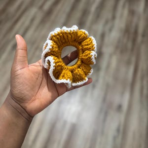 May include: A handmade scrunchie in a sunflower design. The scrunchie is crocheted with a mustard yellow yarn and has a white ruffled edge. This hair accessory adds a touch of handmade charm to any hairstyle.