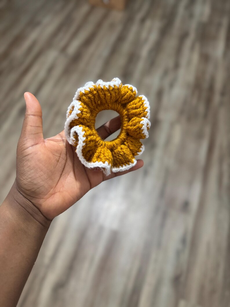 May include: A handmade scrunchie in a sunflower design. The scrunchie is crocheted with a mustard yellow yarn and has a white ruffled edge. This hair accessory adds a touch of handmade charm to any hairstyle.