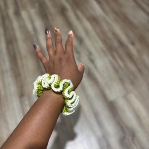 May include: A handmade crochet scrunchie in a wavy pattern. The scrunchie features a combination of white and light green yarn. The accessory is designed to be worn around the wrist or in the hair. The scrunchie is a stylish hair accessory.