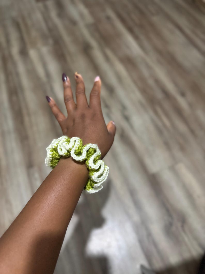 May include: A handmade crochet scrunchie in a wavy pattern. The scrunchie features a combination of white and light green yarn. The accessory is designed to be worn around the wrist or in the hair. The scrunchie is a stylish hair accessory.