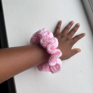 May include: A pink and white crocheted scrunchie. The scrunchie is made of soft yarn and is wrapped around a wrist. The scrunchie is a light pink color with white accents. The background is a white surface.