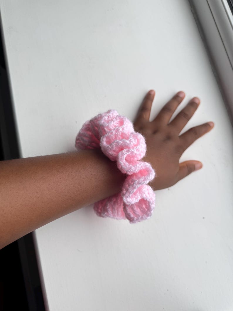 May include: A pink and white crocheted scrunchie. The scrunchie is made of soft yarn and is wrapped around a wrist. The scrunchie is a light pink color with white accents. The background is a white surface.