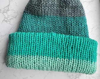 May include: A hand-knitted beanie hat in shades of teal, turquoise, and gray. The hat features a folded brim and a ribbed knit pattern. The top section transitions from gray to teal, while the brim is a lighter shade of teal.