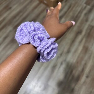 May include: A light purple, handmade crochet scrunchie. The scrunchie is made of soft yarn and has a ruffled design. The scrunchie is worn on a wrist. The scrunchie is a hair accessory.