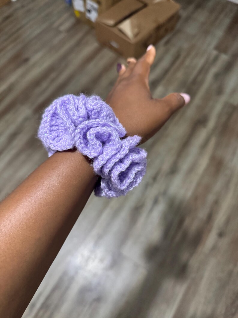 May include: A light purple, handmade crochet scrunchie. The scrunchie is made of soft yarn and has a ruffled design. The scrunchie is worn on a wrist. The scrunchie is a hair accessory.