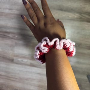 May include: A handmade crochet scrunchie in red and light pink. The scrunchie is worn on a wrist and has a ruffled design. The yarn appears to be soft and the colors are vibrant. This accessory is perfect for adding a pop of color to any outfit.