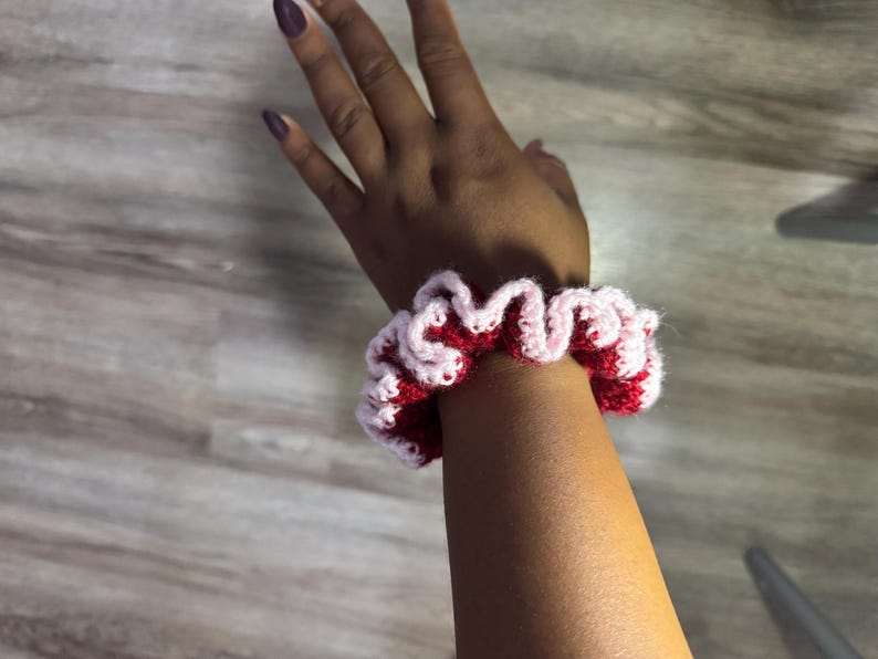 May include: A handmade crochet scrunchie in red and light pink. The scrunchie is worn on a wrist and has a ruffled design. The yarn appears to be soft and the colors are vibrant. This accessory is perfect for adding a pop of color to any outfit.