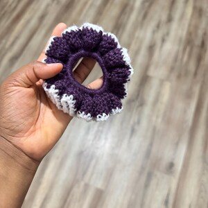 May include: A handmade scrunchie in a deep purple color with a white scalloped edge. The scrunchie is made of a soft, textured yarn and is held in a hand. This hair accessory is perfect for adding a touch of color and style to any hairstyle.
