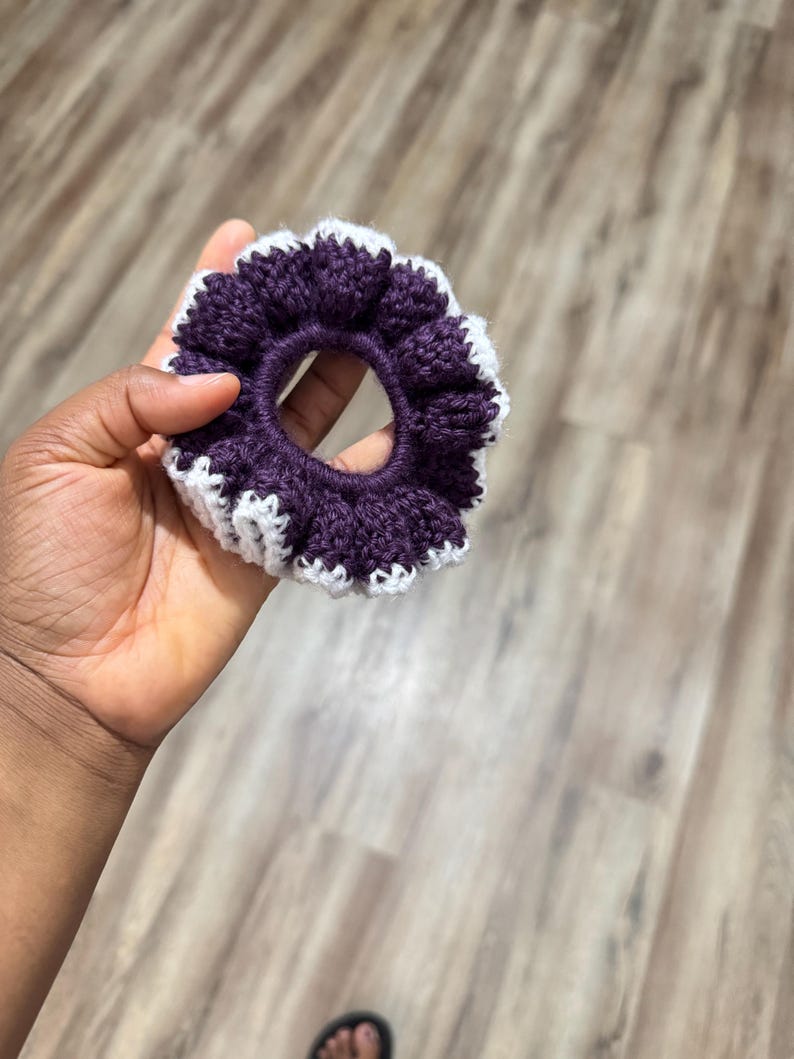 May include: A handmade scrunchie in a deep purple color with a white scalloped edge. The scrunchie is made of a soft, textured yarn and is held in a hand. This hair accessory is perfect for adding a touch of color and style to any hairstyle.