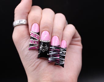 May include: A hand displaying long, artificial nails. The nails feature a pink base with black and decorative designs. One nail has a zebra stripe pattern, another has a heart design, and others have rhinestones.