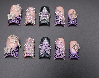 Puede incluir: Un conjunto de diez uñas postizas con varios diseños. Algunas uñas presentan un estampado de leopardo rosa y morado, mientras que otras tienen adornos de estrellas y cuentas plateadas. Las uñas están dispuestas sobre una superficie gris.