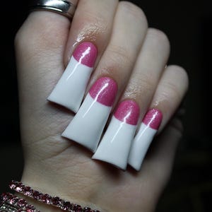 May include: Close-up of a hand with long, white and pink nail art. The nails are a square shape with a white base and a pink glitter tip. A silver ring and a pink bracelet are also visible.