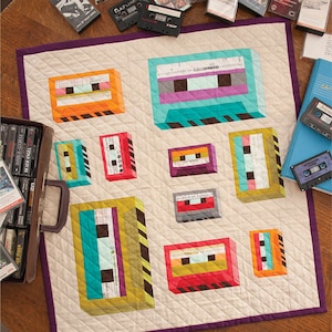 May include: A quilted throw blanket featuring colorful cassette tape designs in various sizes. The tapes are in shades of orange, teal, red, and purple, set against a cream background with a purple border. The blanket is quilted with a diamond pattern.