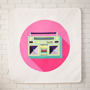 Peut inclure: Une courtepointe blanche avec un grand cercle rose représentant un motif de boombox rétro. Le boombox est turquoise, jaune et violet avec une longue ombre. La courtepointe a un motif matelassé.