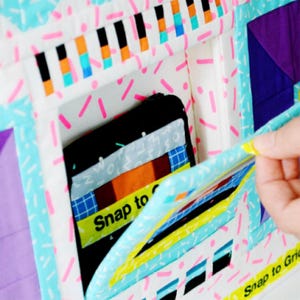 May include: A quilted fabric panel featuring a white background with pink sprinkles and geometric patterns in purple, orange, and blue. A black fabric insert is visible with a yellow label reading Snap to Grid, and a flap with matching text.