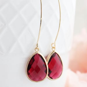 May include: Gold-tone drop earrings featuring faceted, teardrop-shaped, deep red gemstones. The earrings are suspended from fine gold wires. The background is a white, textured surface with a soft pink floral element.