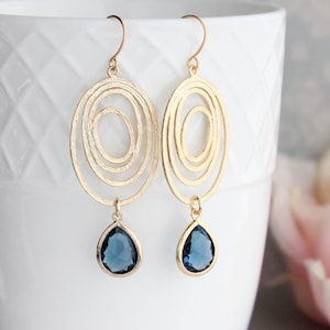 May include: Gold earrings with a teardrop shaped blue gemstone dangling from a circular design with multiple concentric circles.