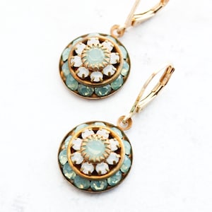 May include: A pair of gold-coloured earrings with a circular design. Each earring features a central pale blue stone surrounded by white and pale blue gemstones. The earrings have a lever-back closure.
