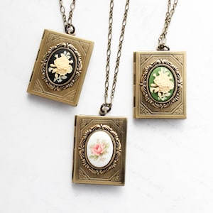 May include: Three rectangular brass-toned metal book-shaped lockets on matching chains. Each locket features an oval cameo with a floral relief. One has a black background, one green, and one white with a pink rose illustration.